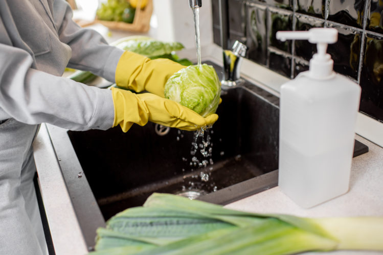 Woman in protective gloves washing fresh vegetables at home on the kitchen, detergent in the foreground. Concept of hygiene during the coronavirus epidemic
